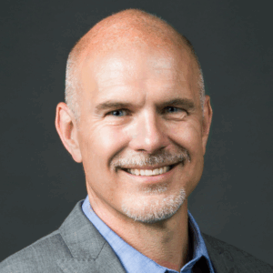 A headshot of a smiling person with a bald head, wearing a suit jacket and a collared shirt against a dark background.