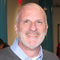 A headshot of a person smiling, with short gray hair and a trimmed beard, wearing a light blue collared shirt and dark sweater.