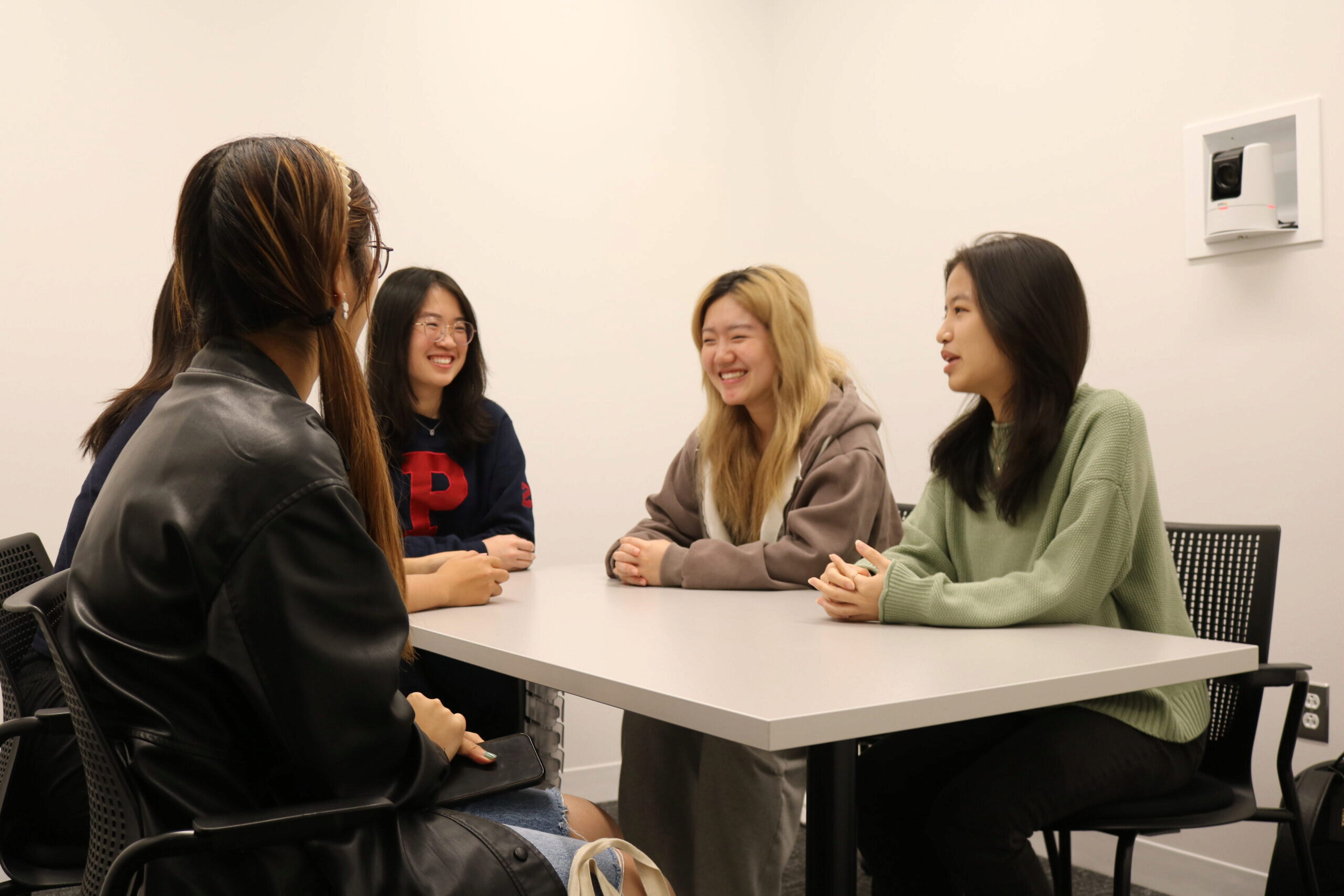 A group of people sitting around a table, engaged in conversation and smiling, in a meeting room.