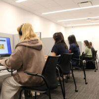 A small group of individuals is seated in a computer lab, each wearing headphones and using desktop computers in separate cubicles.