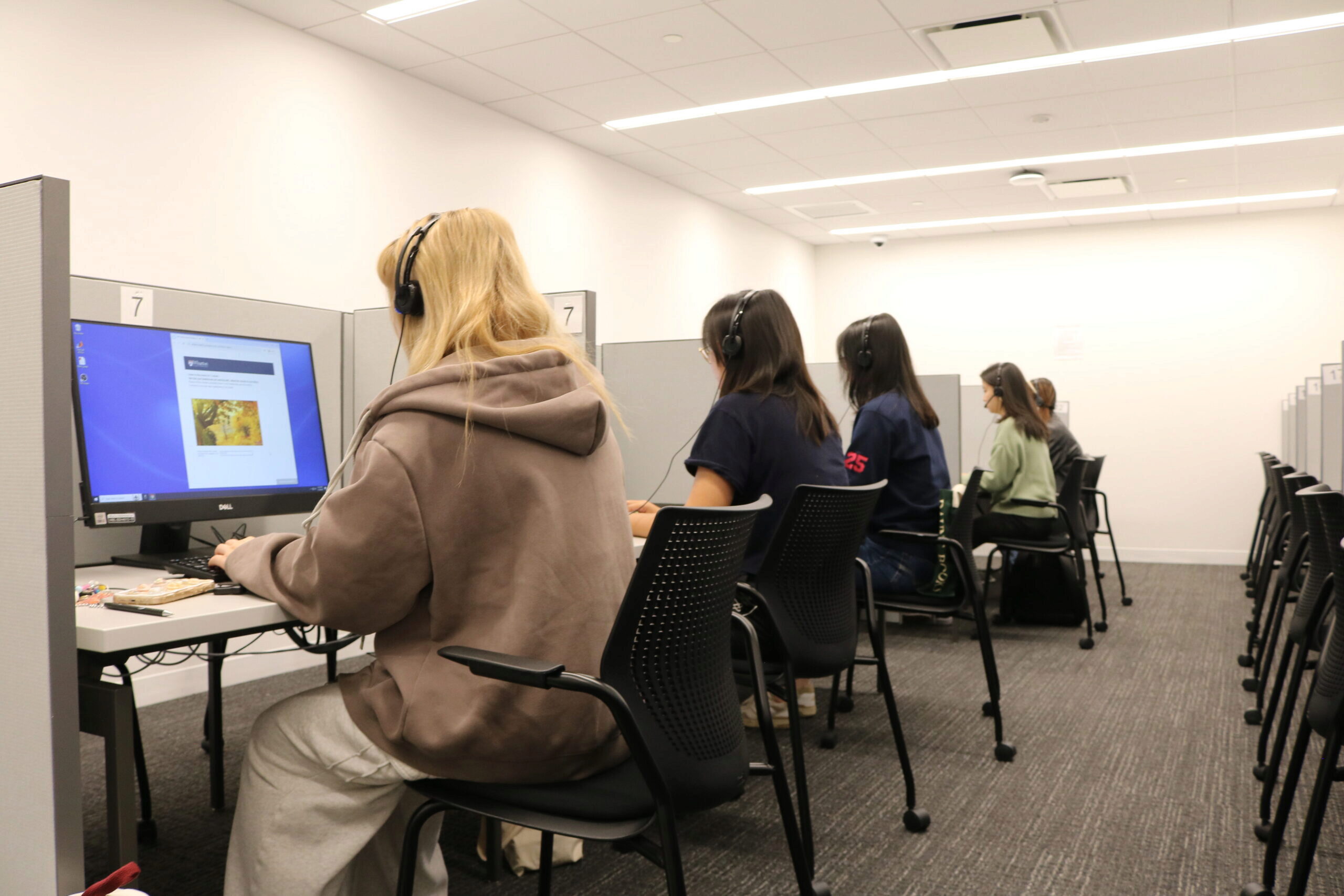 A small group of individuals is seated in a computer lab, each wearing headphones and using desktop computers in separate cubicles.