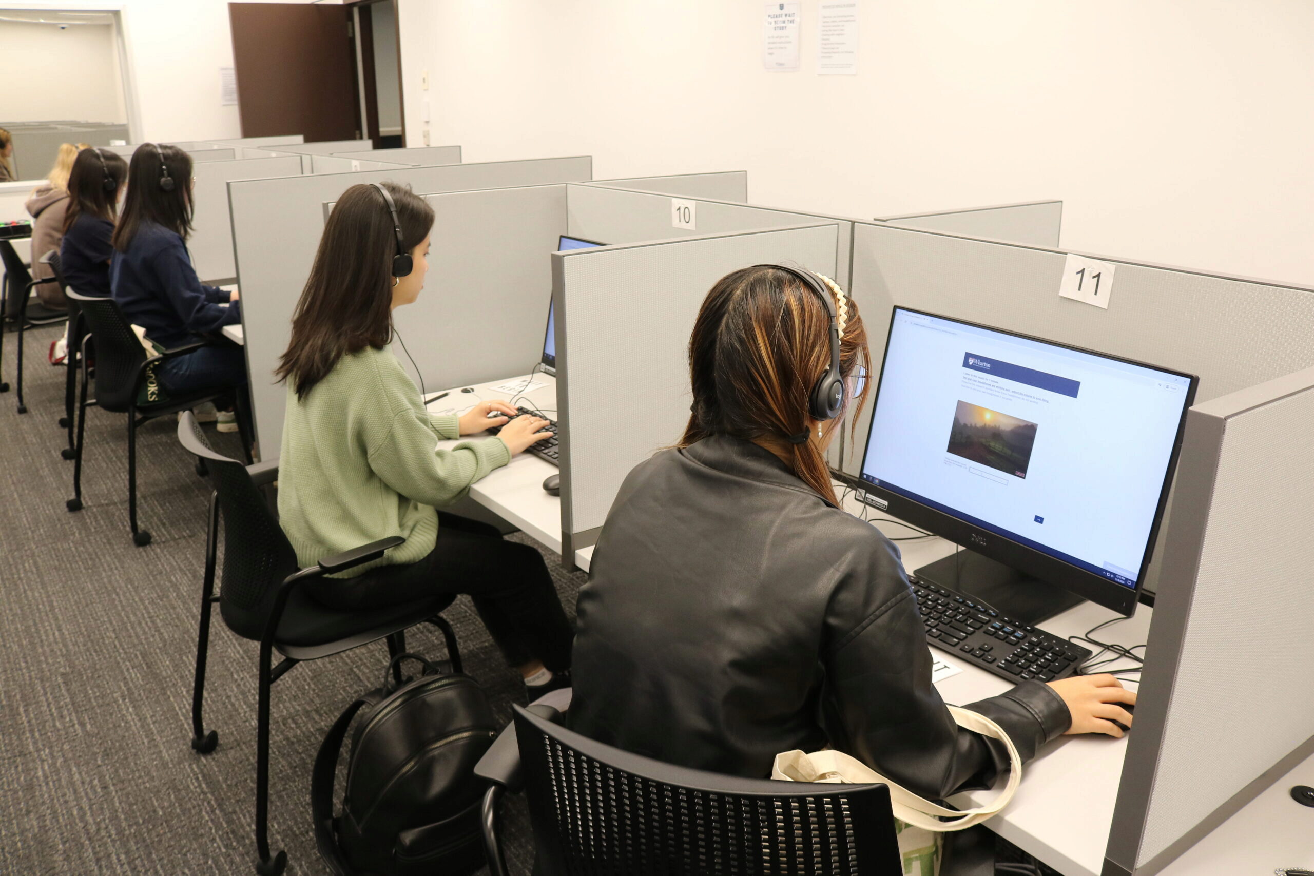 A group of people in a computer lab using monitored workstations, each equipped with headphones, focused on their screens in a quiet environment.