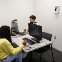 Two people sitting at a table in a focus room with computer monitors and microphones. The room number "407" is displayed on the wall.