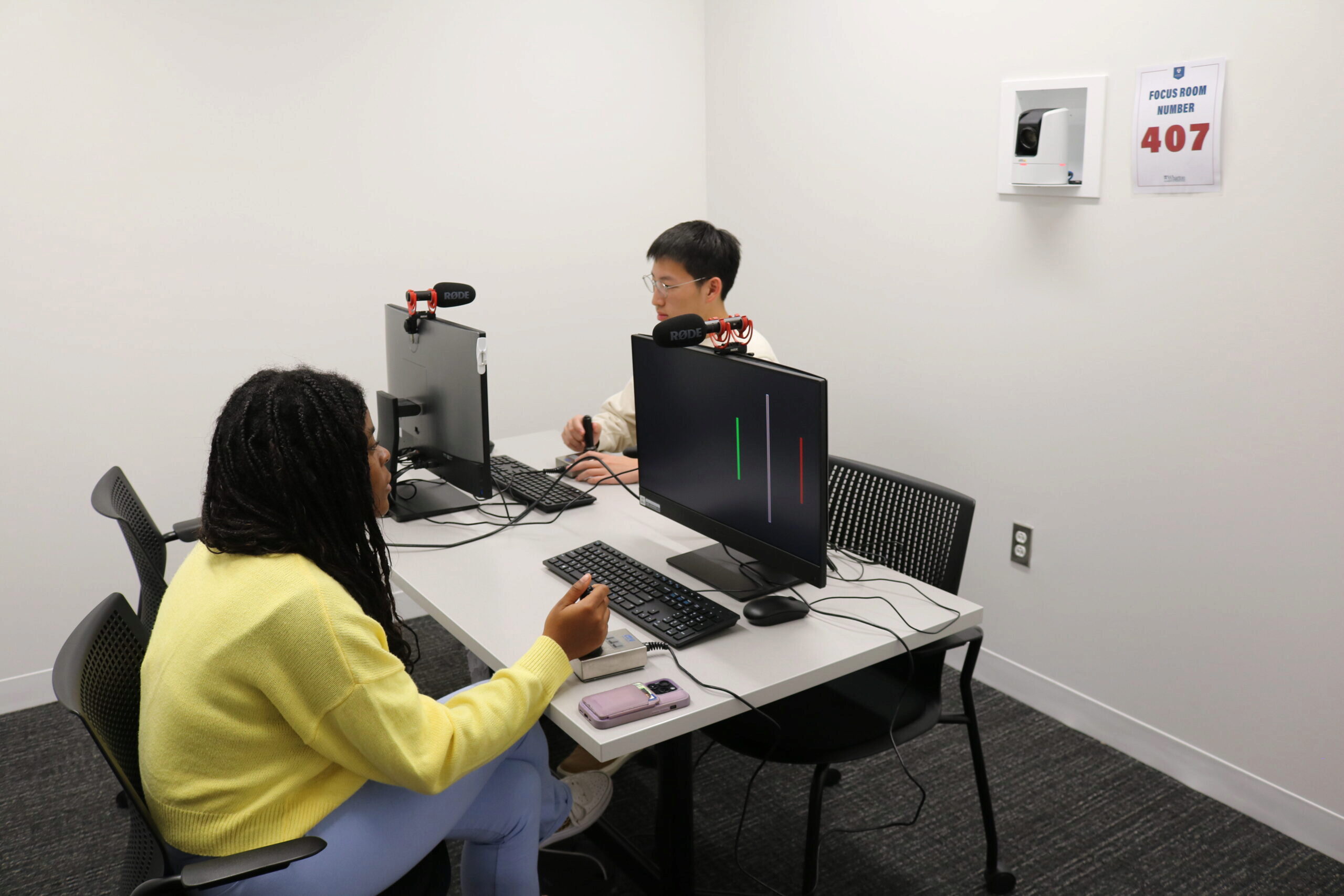 Two people sitting at a table in a focus room with computer monitors and microphones. The room number "407" is displayed on the wall.