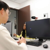 Two people in a room with a computer and microphone setup. One person is sitting at the desk, looking at the monitor, with a microphone placed on top. The scene suggests a recording or communication setting.