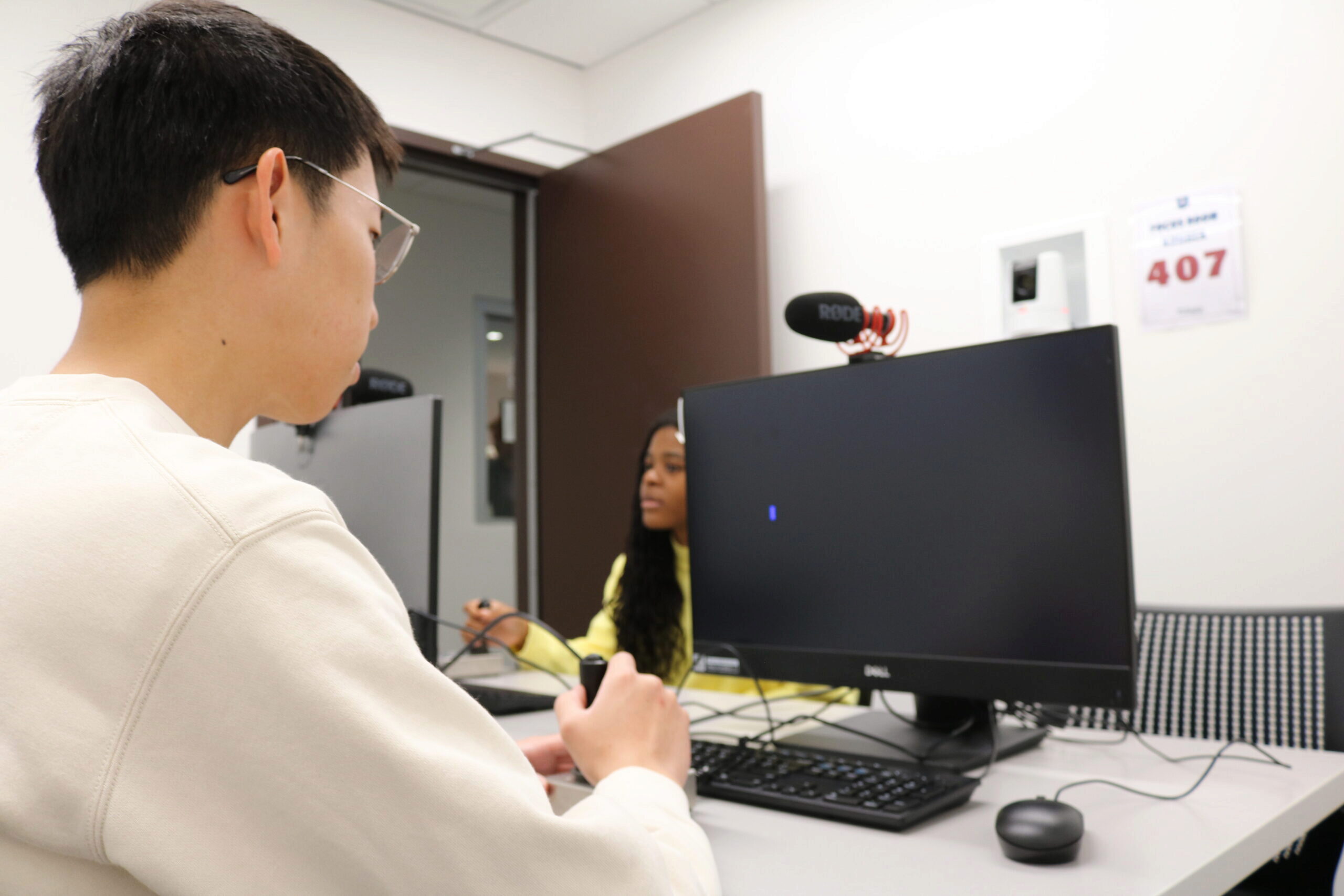 Two people in a room with a computer and microphone setup. One person is sitting at the desk, looking at the monitor, with a microphone placed on top. The scene suggests a recording or communication setting.