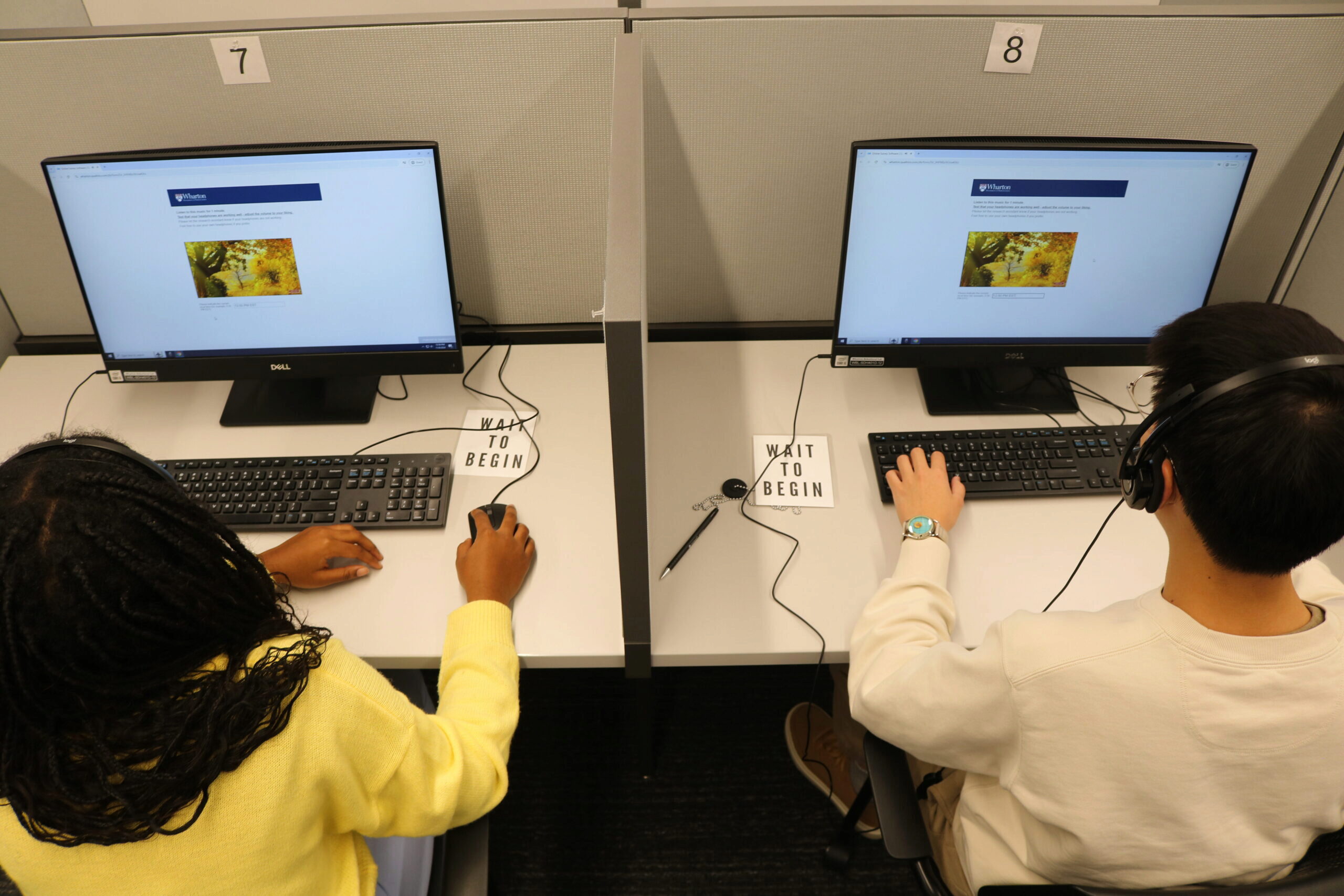 Two people wearing headphones seated at computer stations in a partitioned cubicle, viewing screens with the text "WAIT TO BEGIN" on the desk.