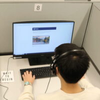 A person wearing headphones is sitting at a computer workstation in a cubicle, looking at a monitor displaying an educational or informational webpage.