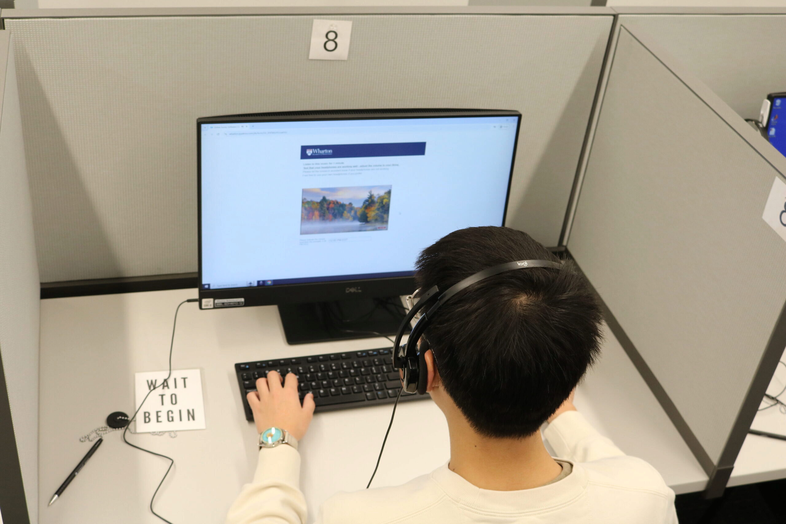 A person wearing headphones is sitting at a computer workstation in a cubicle, looking at a monitor displaying an educational or informational webpage.