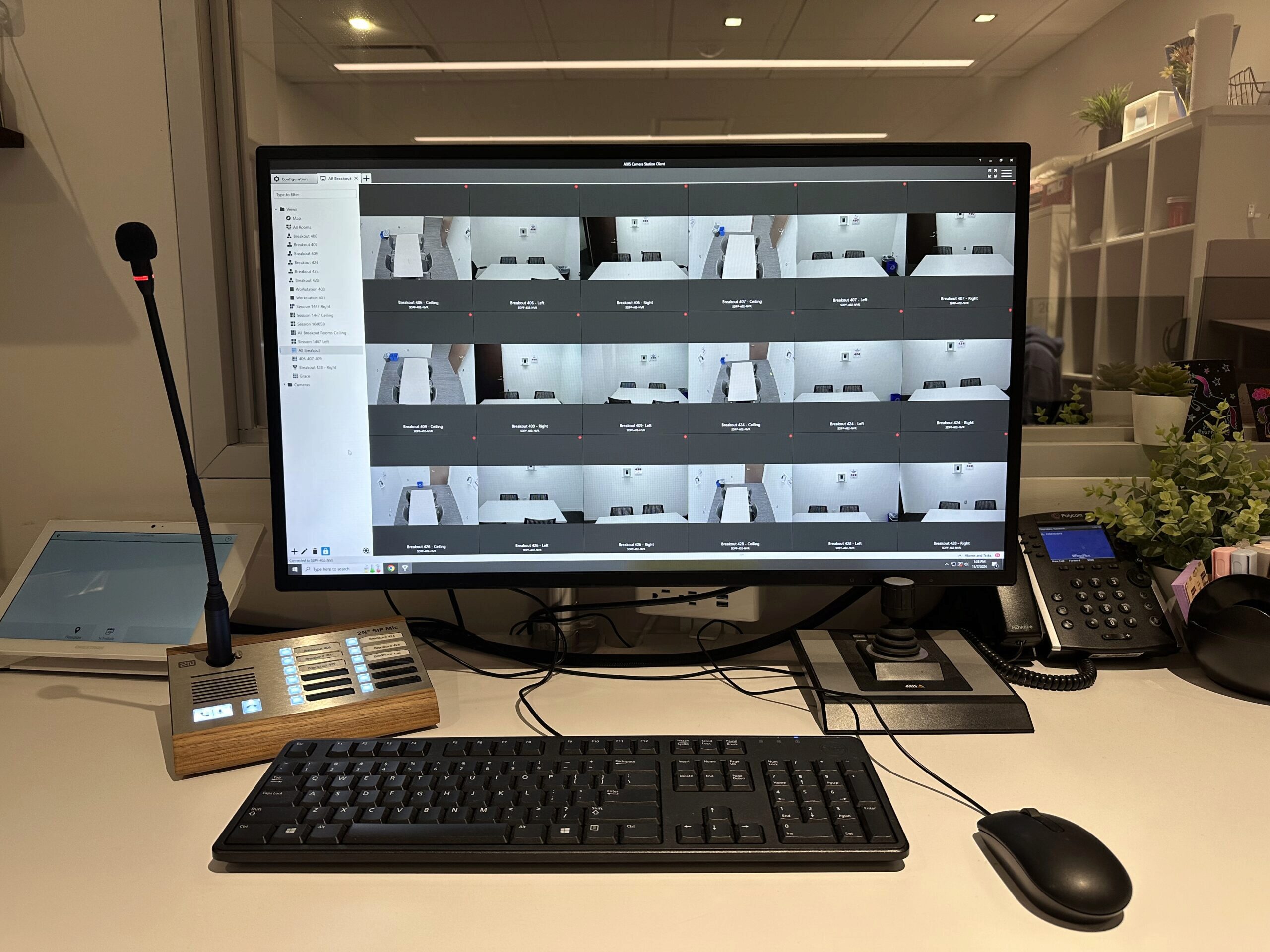 A desk with a computer monitor displaying multiple security camera feeds. On the desk are a keyboard, mouse, microphone, and various electronic equipment. There are also potted plants and a telephone.