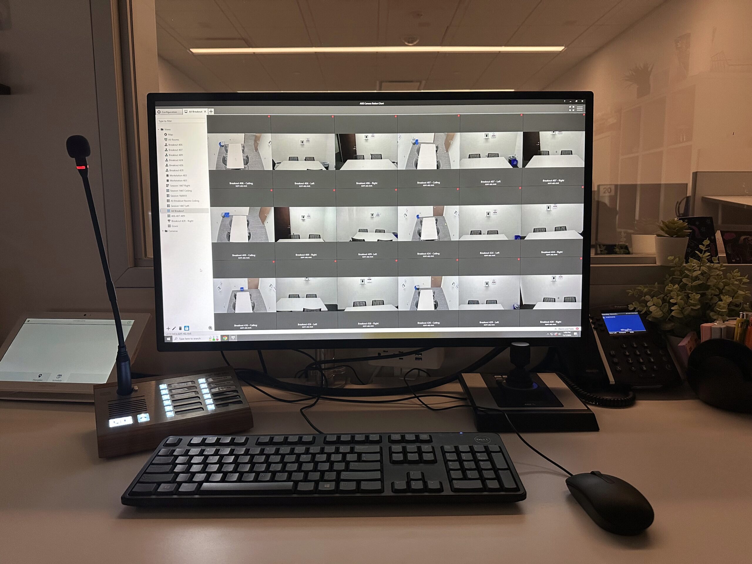 A security control room setup with a computer monitor displaying multiple surveillance camera feeds, a keyboard, mouse, microphone, and communication device on a desk.