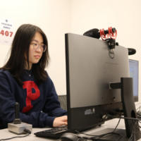 A person working at a computer station in a focus room, wearing glasses and a dark sweatshirt with a red letter "P." A microphone is mounted on the monitor.