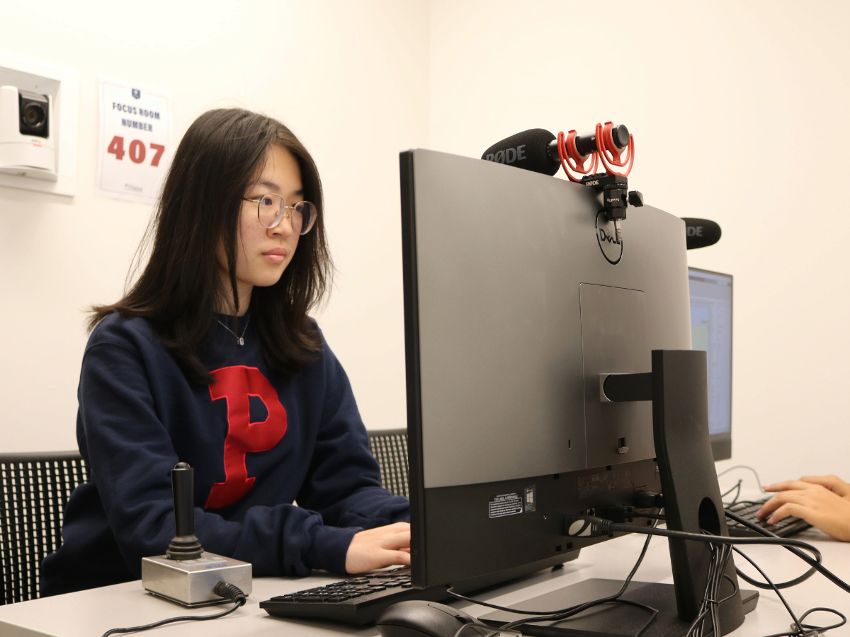 A person working at a computer station in a focus room, wearing glasses and a dark sweatshirt with a red letter "P." A microphone is mounted on the monitor.