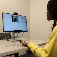 A person in a yellow sweater sits at a table, interacting with a device that measures physiological data. A monitor displays their video feed along with data graphs. A focus room sign is visible on the wall.