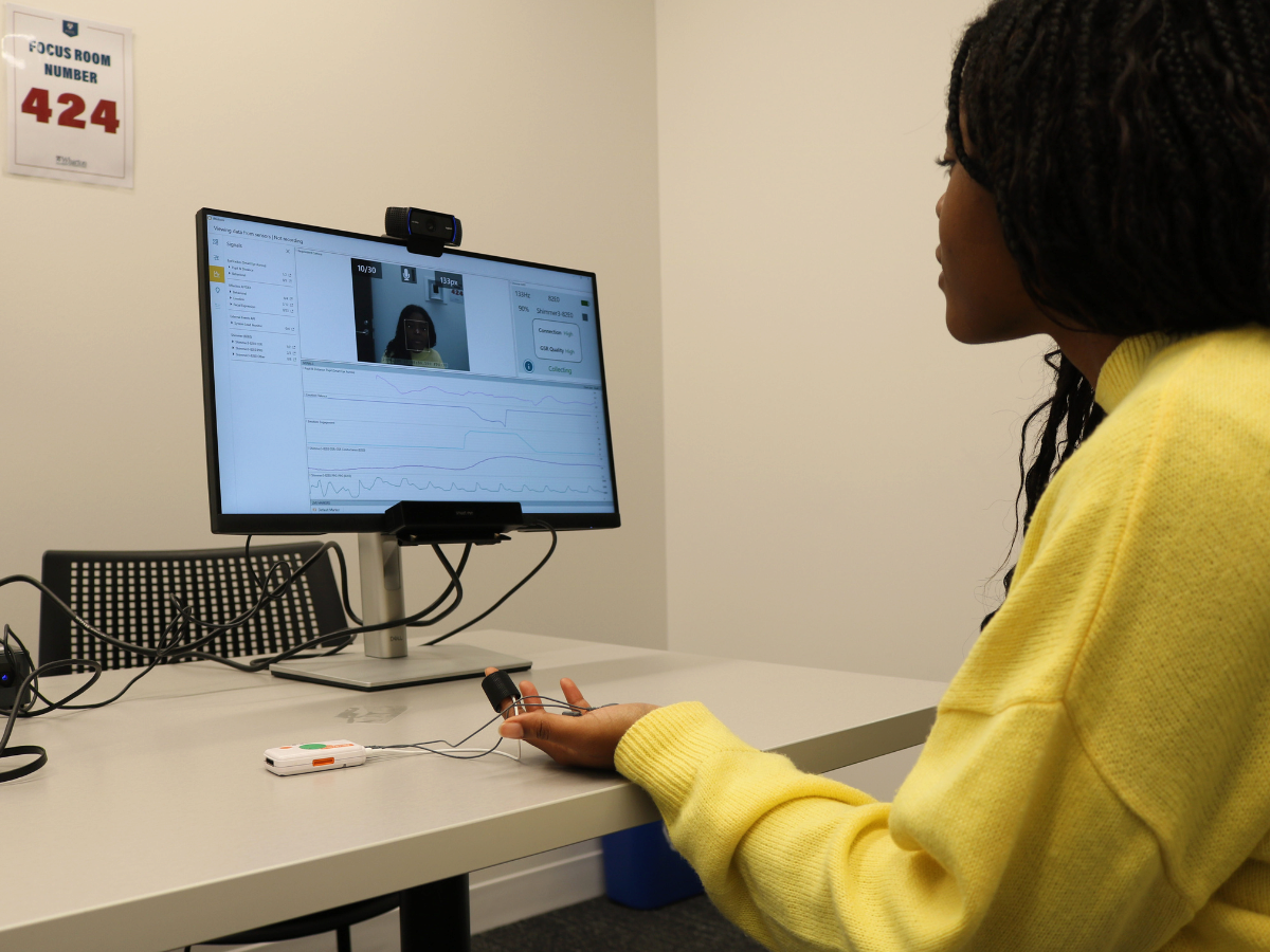 A person in a yellow sweater sits at a table, interacting with a device that measures physiological data. A monitor displays their video feed along with data graphs. A focus room sign is visible on the wall.