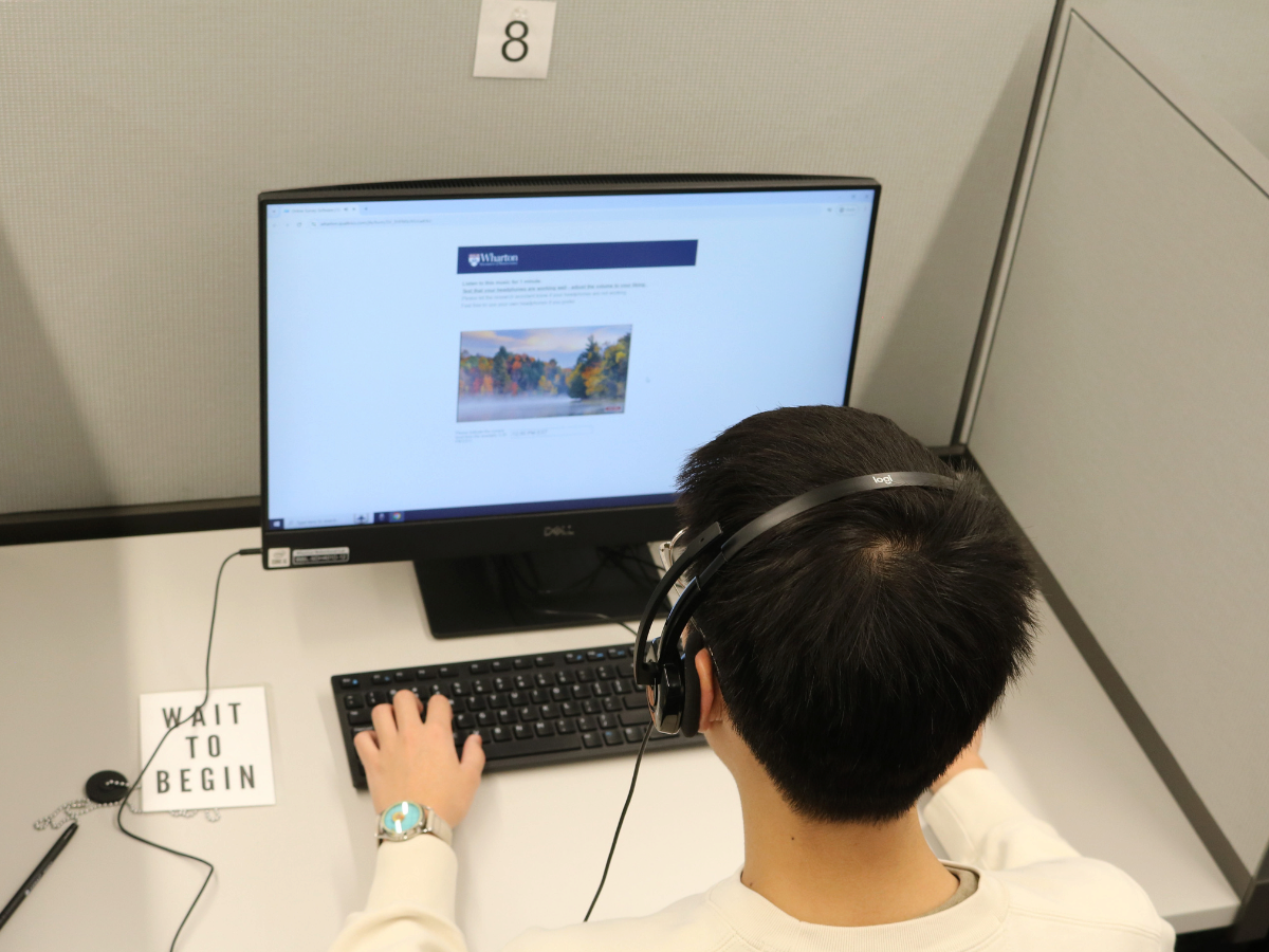 A person wearing headphones sits at a desk using a computer. The monitor displays a webpage, and a sign on the desk says "WAIT TO BEGIN." The setting appears to be a study or work cubicle.