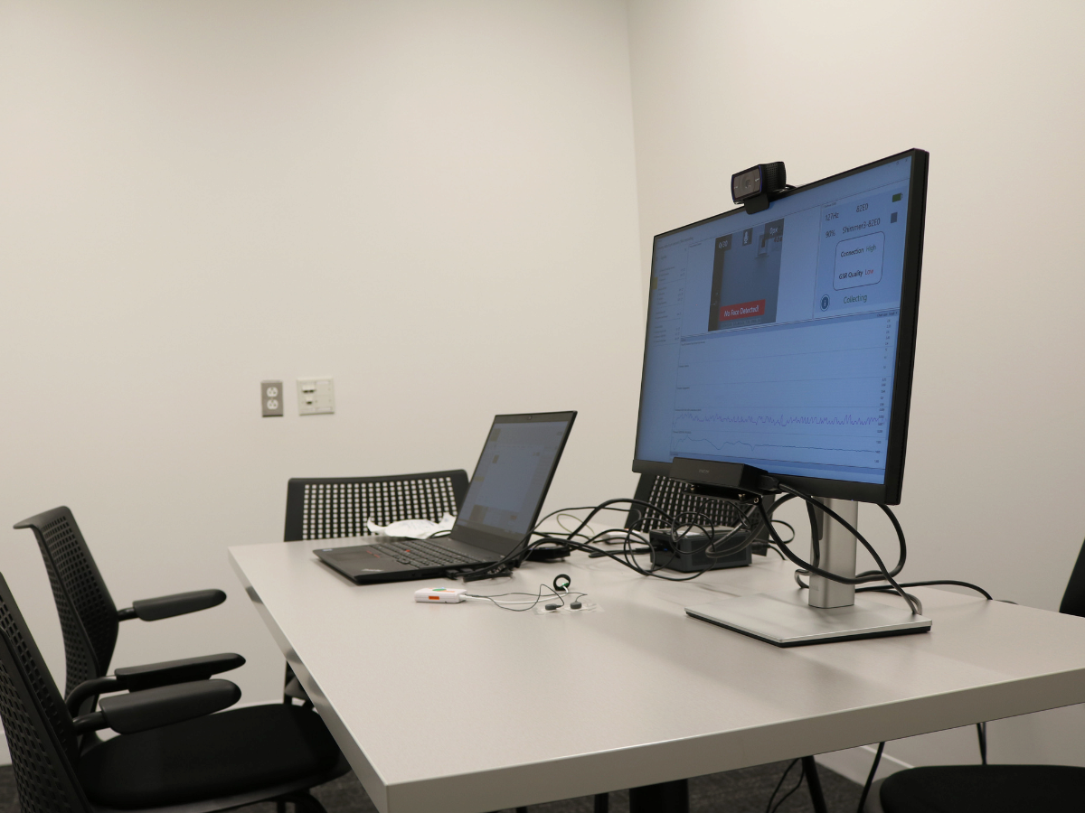 A small meeting room with a rectangular table, two chairs, a laptop, a monitor displaying data, and a webcam.