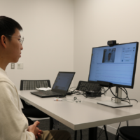 A person sits at a table in an office setting, facing a computer monitor displaying a video call or online test. A laptop is on the table, suggesting a digital or remote interaction.