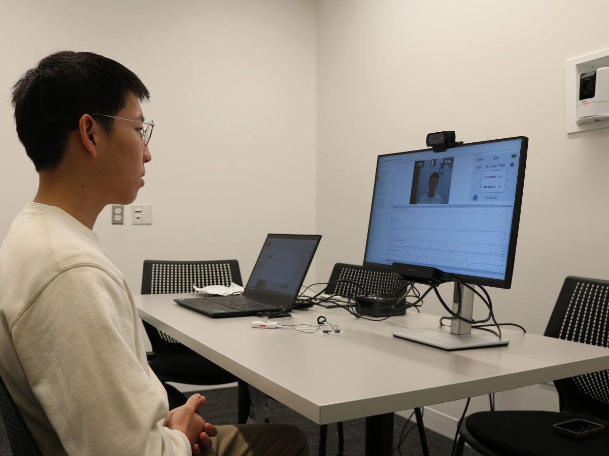 A person sits at a table in an office setting, facing a computer monitor displaying a video call or online test. A laptop is on the table, suggesting a digital or remote interaction.