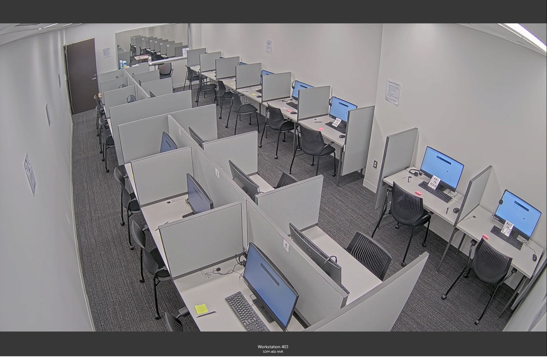 Image of a computer lab or testing center with individual workstations separated by gray dividers. Each station has a monitor, keyboard, and chair.