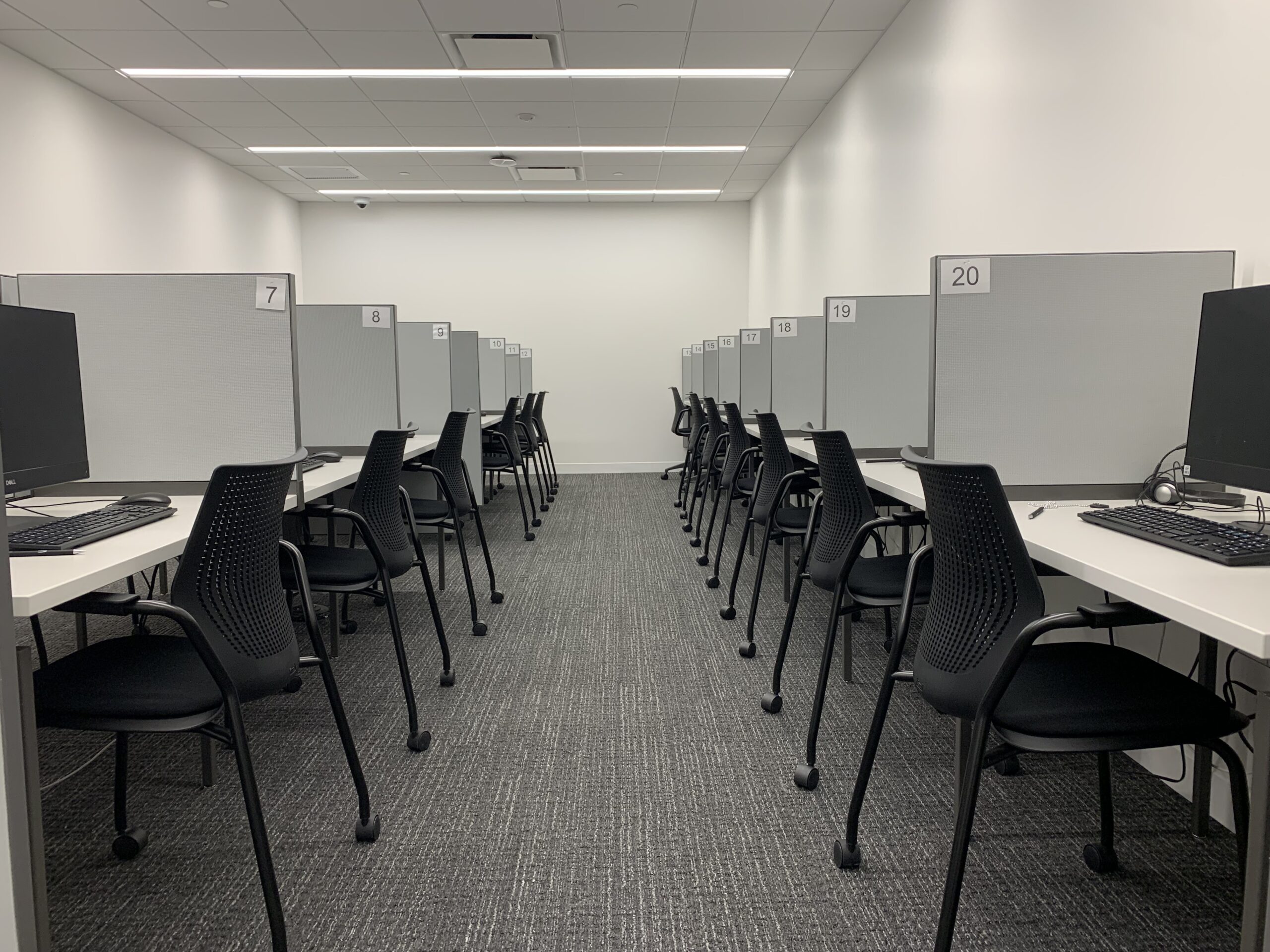 A modern computer lab with cubicles, each equipped with a computer monitor, keyboard, and chair. The setup suggests it is used for individual work or testing.