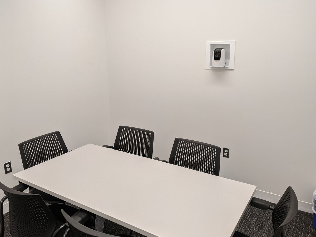 A small conference room with a rectangular table and several black chairs. A camera is mounted on the wall, and there are electrical outlets nearby.