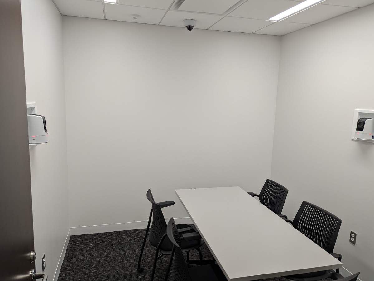 A small, minimalist conference or meeting room with a table and several chairs. The room has white walls and some mounted equipment, such as cameras, possibly for security or video conferencing.