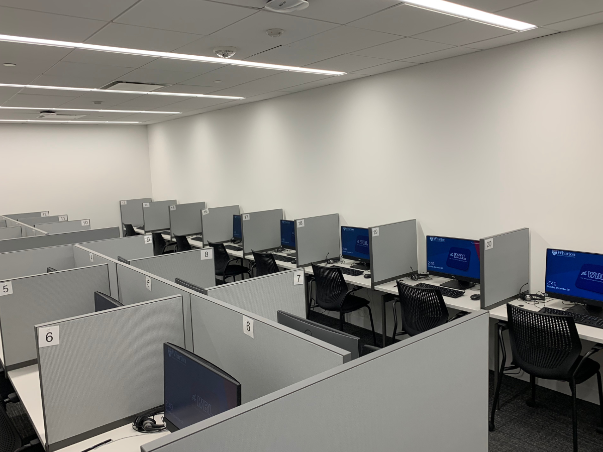 A modern computer lab with numbered workstations, each equipped with a computer, chair, and a partition for privacy. The room has a clean, neutral design.