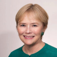 Headshot of a smiling person with short, light hair, wearing a teal top and small earrings, against a neutral background.