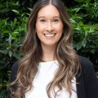 Headshot of a person with long, wavy hair smiling in front of lush green foliage, wearing a white top and a dark jacket.
