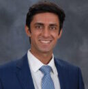 Headshot of person wearing a suit and tie, smiling against a gray background.