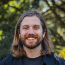 Headshot of a person with long hair and a beard, smiling in an outdoor setting with blurred trees in the background.
