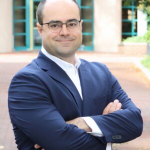 Headshot of a person wearing glasses and a dark suit, standing confidently with arms crossed outdoors.