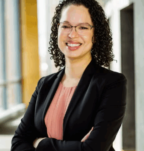 Headshot of person with curly hair, wearing glasses, a black blazer, and a peach blouse, smiling with arms crossed in a well-lit hallway.