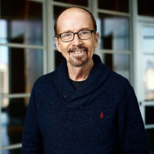 Headshot of a smiling person with glasses, wearing a dark blue sweater, set against a background of windows.