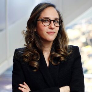 Headshot of person wearing glasses and a black outfit, with wavy hair and a confident expression.